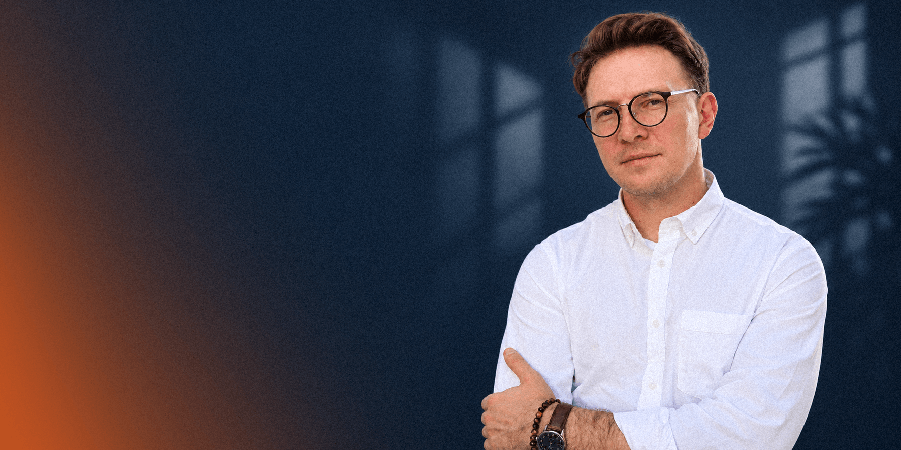 Marketing strategist in a white shirt and glasses, arms crossed, against a dark blue wall.