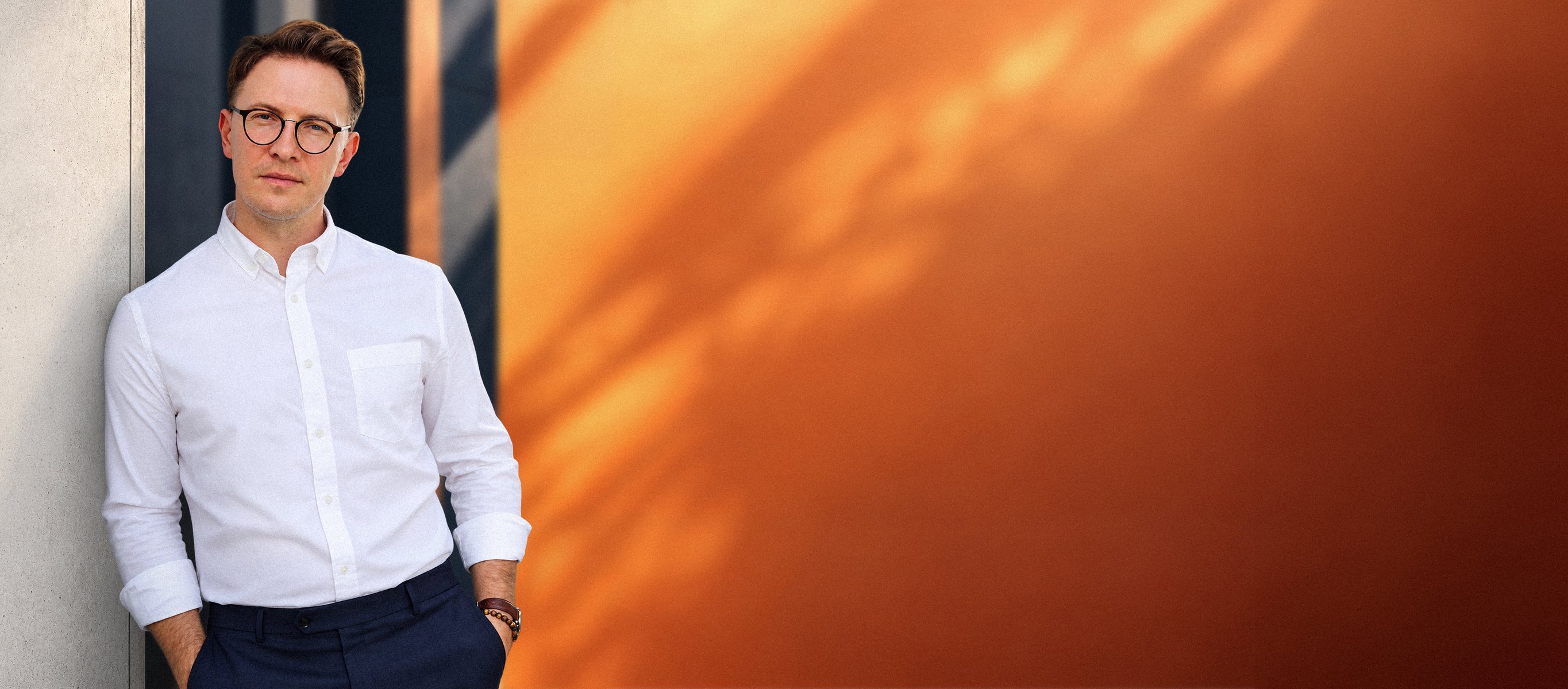Marketing strategist in a white shirt and glasses leaning against a concrete wall beside a sunlit orange facade.